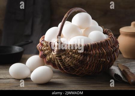 Wicker basket full of white chicken eggs on wooden table. Frying pan and clay pot on background. Stock Photo