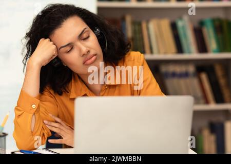 Tired mixed race female student sleeping on table in front of laptop computer, preparing for exams in library Stock Photo