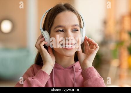 Portrait of happy caucasian teenage girl enjoying music touching wireless headphones and smiling, resting at home Stock Photo