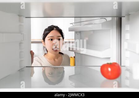 Upset chinese woman looking at lonely tomato in her fridge Stock Photo