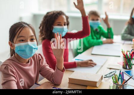 Diverse School Children Wearing Face Masks Raising Arms In Classroom Stock Photo