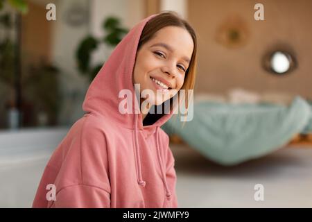 Portrait of cheerful caucasian teen girl wearing hood and smiling at camera, posing sitting in bedroom interior at home Stock Photo