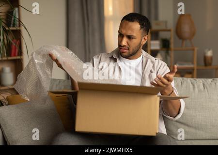 Dissatisfied black male customer opening box from online store, taking out packaging, unhappy with received item at home Stock Photo
