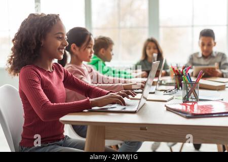 Black Schoolgirl Using Laptop Sitting With Multiethnic Classmates In Classroom Stock Photo