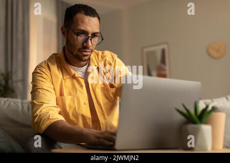Focused millennial black man in glasses using laptop, working online, having online business meeting at home office Stock Photo
