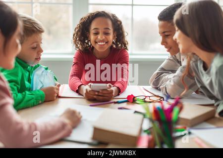 Happy African Schoolgirl Using Phone Sitting With Classmates In Classroom Stock Photo