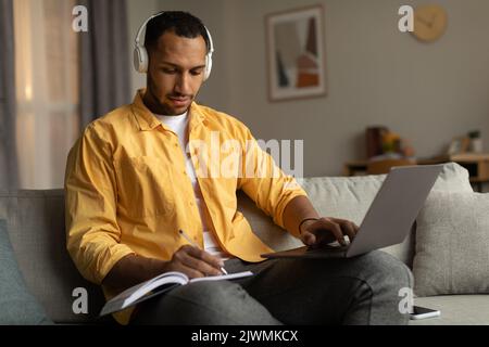 Focused young black man in headphones having online business conference on laptop, taking notes at home Stock Photo