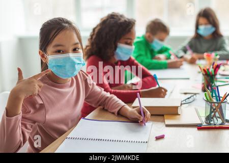 Japanese Schoolgirl Wearing Face Mask Gesturing Thumbs Up In Classroom Stock Photo