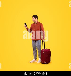 Traveler Man With Suitcase Using Cellphone Texting On Yellow Background Stock Photo
