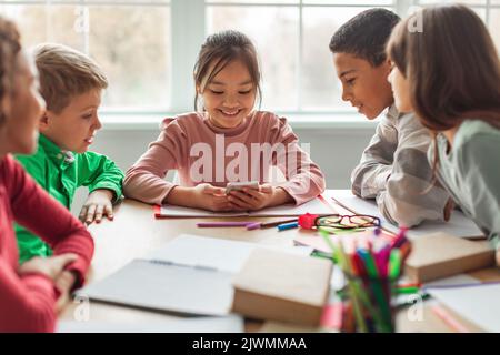 Diverse School Kids Using Cellphone During Break In Classroom Stock Photo