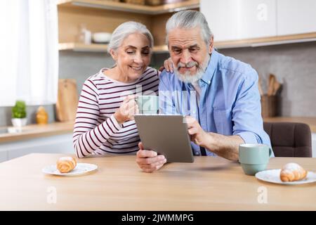 Happy Senior Spouses Using Digital Tablet Together While Having Breakfast In Kitchen Stock Photo