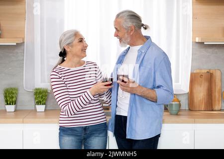 Happy Senior Spouses Drinking Wine And Chatting Together In Kitchen Interior Stock Photo