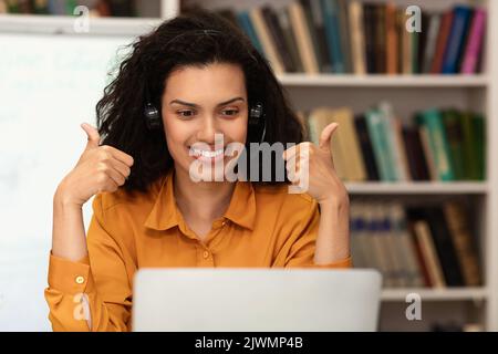 Happy female teacher giving lesson on web using laptop and gesturing thumbs up at webcamera, sitting in library Stock Photo
