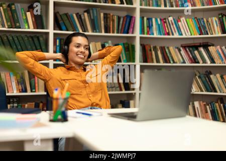 Relaxed mixed race female student sitting with hands behind head near laptop, having break after online class in library Stock Photo