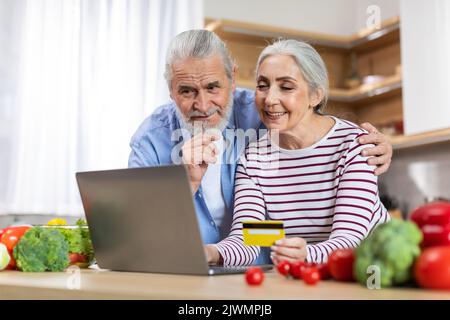 Senior Couple Using Laptop And Credit Card In Kitchen, Ordering Groceries Online Stock Photo