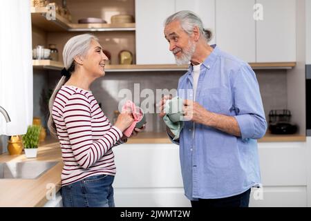 Happy Senior Spouses Sharing Domestic Chores, Wiping Dishes Together In Kitchen Stock Photo