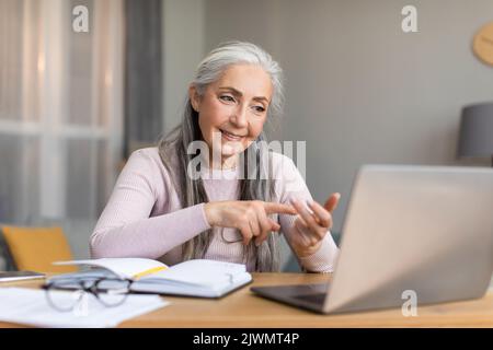 Glad friendly caucasian senior gray-haired female teacher speaks by laptop with student in living room Stock Photo