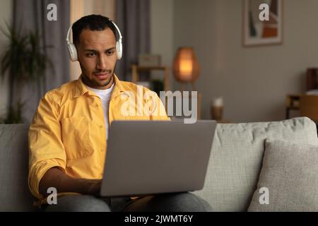 Attractive young black man in headset using laptop computer, having online job, sitting on couch at home, blank space Stock Photo