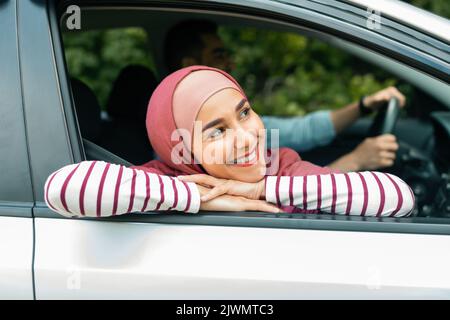 Happy glad young muslim man driving car with husband in hijab, lady looking out the window and enjoying car journey Stock Photo