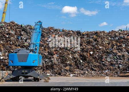 A large pile of scrap metal and waste at a recycling plant with a blue excavator in front Stock Photo