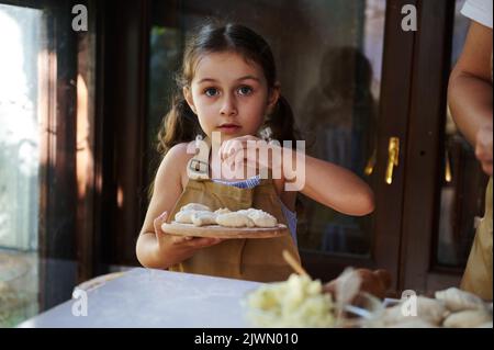 Beautiful little girl in a chef's apron sprinkling flour on a wooden board with homemade dumplings, cooking with her mom Stock Photo