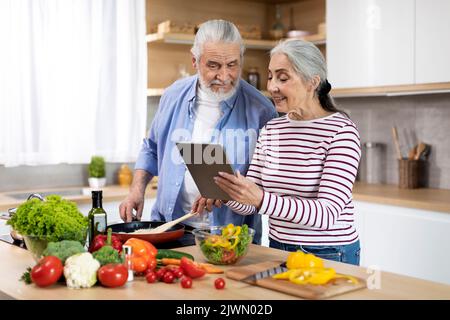 Happy Elderly Spouses Cooking Food In Kitchen And Using Digital Tablet Together Stock Photo