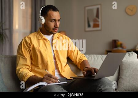 Young black man in headphones having online business conference or webinar on laptop, taking notes at home Stock Photo