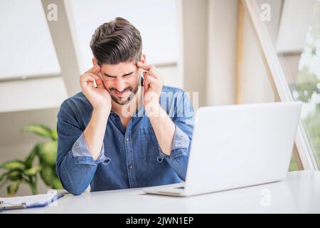 Shot of stressed businessman sitting at desk in office with laptop. Professional man rubbing forehead while suffering from migraine. Stock Photo