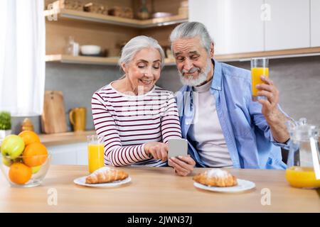 Happy Senior Couple Using Smartphone In Kitchen While Having Breakfast Together Stock Photo