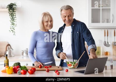 Loving senior spouses cooking together at home, using laptop Stock Photo