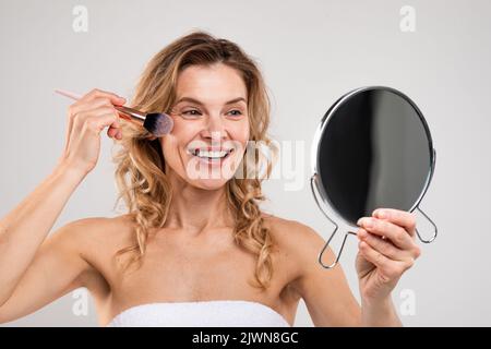Happy Middle Aged Female Holding Mirror And Applying Blush On Face Stock Photo