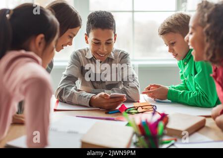 Excited Black Schoolboy Using Phone With Classmates In Modern Classroom Stock Photo