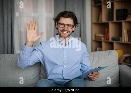 Glad cheerful young caucasian male doctor waving hand at camera in office interior. Online medical support Stock Photo