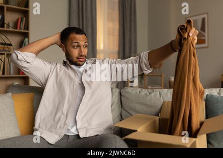 Dissatisfied black male opening box from online shop and looking at wrong delivered clothes, unhappy with received item Stock Photo