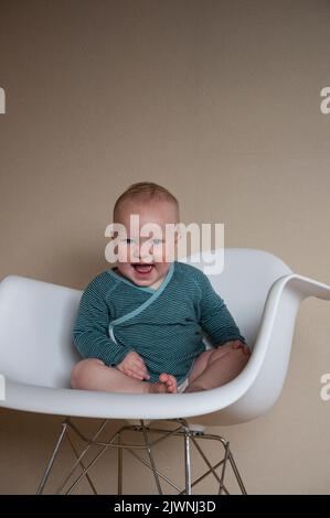 A baby in a striped blue top sits in an Eames-style rocking chair against a neutral background. Stock Photo