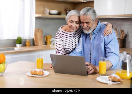Happy elderly spouses spending time with laptop in kitchen Stock Photo