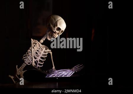 A skeleton sits at a table near a laptop in an apartment in the dark, online work, overwork and stress, a programmer Stock Photo