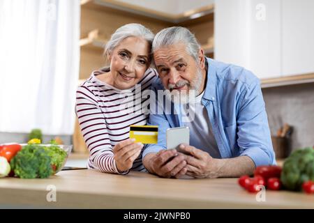 Portrait Of Happy Senior Spouses With Smartphone And Credit Card In Kitchen Stock Photo