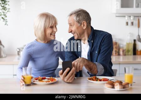 Smiling senior spouses having breakfast and using cell phone Stock Photo