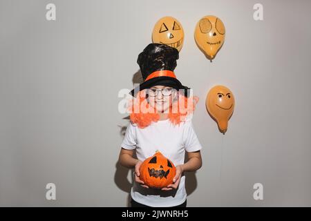 Happy Halloween. Child in orange wig and black hat is holding pumpkin with painted face. The girl is smiling and wearing glasses. Autumn, cheerful moo Stock Photo