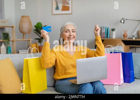 Glad happy caucasian old gray-haired woman sitting on sofa with packages and making victory gesture with laptop Stock Photo