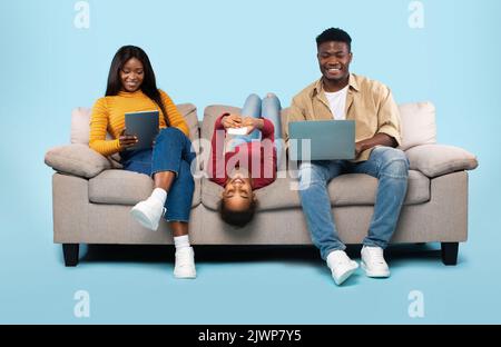 Gadgets addiction concept. African american family of three using electronic devices, sitting on sofa on blue background Stock Photo