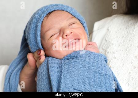 infant newborn boy swaddled in blue towel with head wrapped resting on little bed flower and leaves Stock Photo