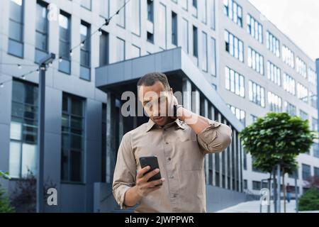 Pensive and excited african american worker outside office building reading bad news, man holding phone. Stock Photo