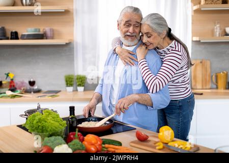Loving Wife Embracing Her Elderly Husband While He Cooking Lunch In Kitchen Stock Photo