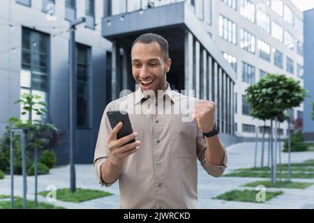 Successful african american businessman outside office building using smartphone, smiling and happy celebrating victory online games, got good news Stock Photo