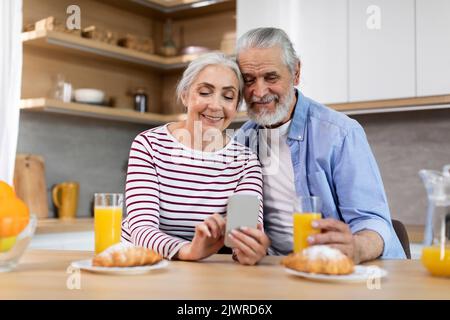 Happy Senior Spouses Spending Time With Smartphone While Having Breakfast In Kitchen Stock Photo