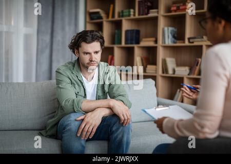 Despaired depressed young caucasian man suffers from mental problems, listens to black female psychologist Stock Photo