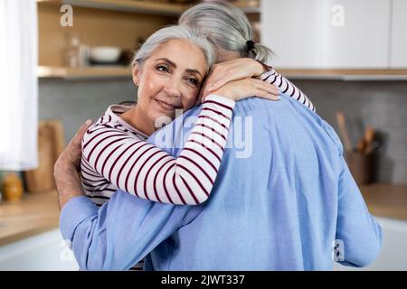 Mature Romance. Beautiful Senior Woman Tenderly Embracing Her Husband In Kitchen Stock Photo