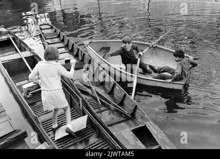 London, England, circa 1967  A young girl steps across moored boats to pass a cup of tea and a bag of sweets to two boys in a rowing boat. They are all members of The Pirate Club. The Pirate Club, a children’s boating club was formed in 1966 at Gilbey’s Wharf on the Regents Canal near Camden, London. Their clubhouse was an old barge and a number of small boats and canoes had been donated for the children’s use. Stock Photo